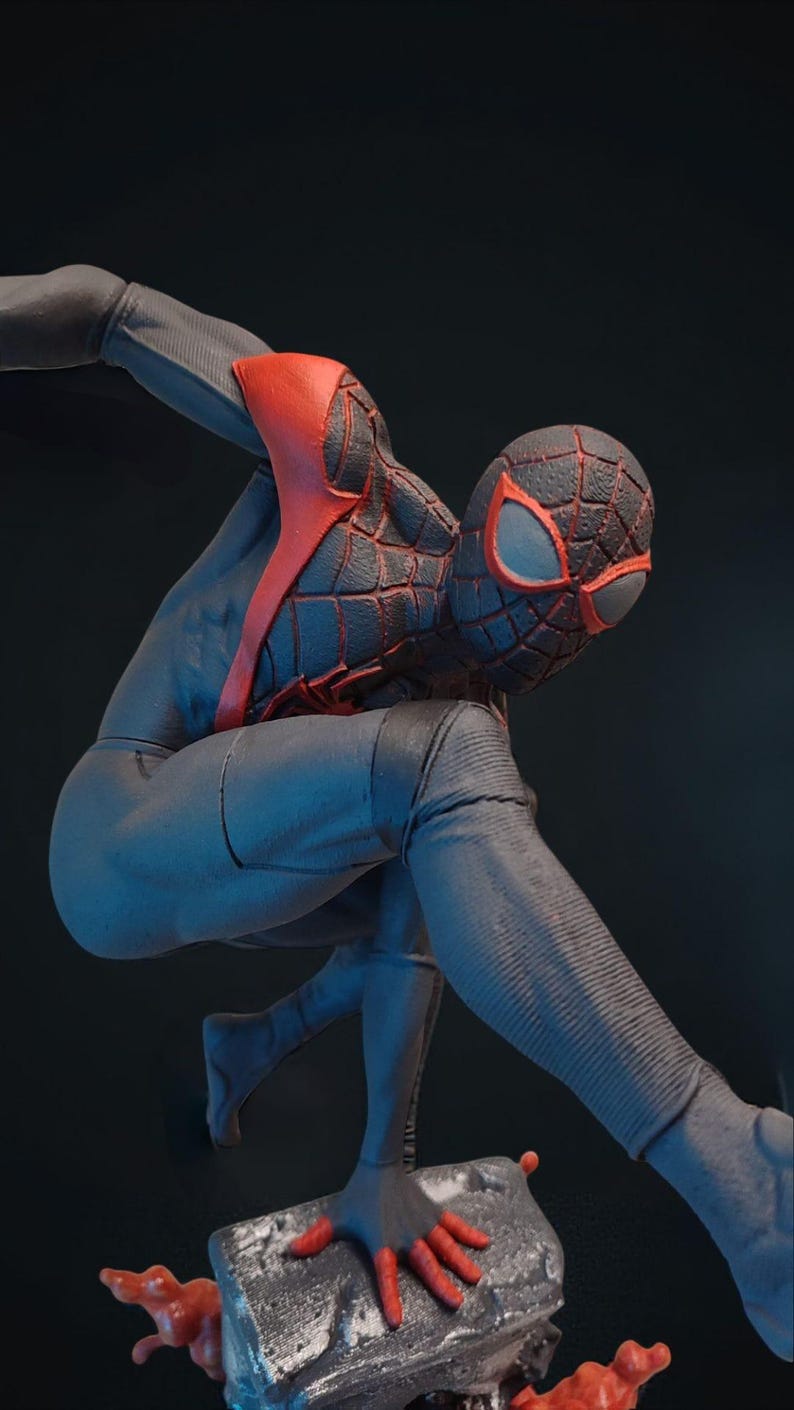 May include: A figurine of a superhero in a dynamic pose. The figure is clad in a black and blue suit with red accents on the chest and mask. The mask features red eye shapes. The figure is posed on a gray base.