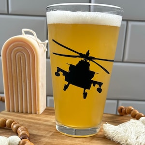 May include: A pint glass filled with beer, featuring a black helicopter silhouette. The glass sits on a wooden tray with a decorative candle and beaded garland with tassels. The background is a tiled wall.