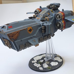 May include: A grey and orange spaceship model with a wolf insignia. The model is on a black base with white rocks and green grass.