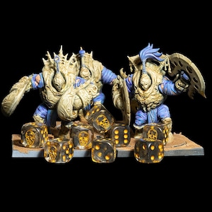 May include: A tabletop gaming scene featuring two large, armored figures in beige and blue, with shields and weapons. In front are several clear dice with gold skull designs. The figures stand on a small, textured base.