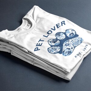 Puede incluir: Camiseta blanca con un diseño de huella de pata floral azul y las palabras "PET LOVER" en azul. El texto "EST. 1978" también está impreso en la camiseta. La camiseta está doblada y apilada sobre una superficie azul oscuro.