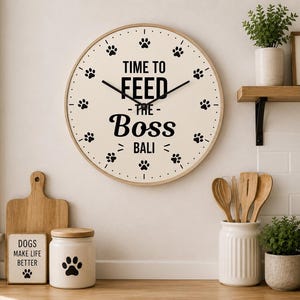 Puede incluir: Un reloj de pared redondo con una esfera clara y un marco de madera. La esfera del reloj presenta huellas de patas negras y el texto "TIME TO FEED THE BOSS BALI". El reloj está rodeado de decoración de cocina.