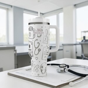 May include: White Stanley tumbler with a handle and a straw, featuring black line art of medical symbols, including a stethoscope, first aid kit, and the text "CARE COMPASSION EVERY CALL". The tumbler is on a white surface with a stethoscope.