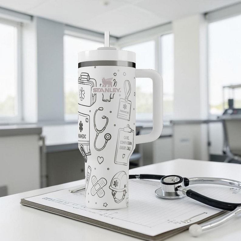 May include: White Stanley tumbler with a handle and a straw, featuring black line art of medical symbols, including a stethoscope, first aid kit, and the text "CARE COMPASSION EVERY CALL". The tumbler is on a white surface with a stethoscope.