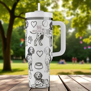 Puede incluir: Vaso térmico blanco con asa y pajita, con un estampado repetido de perros dálmatas blancos y negros, corazones, huellas de patas y cuencos para perros. El logotipo de Stanley es visible. El vaso está sobre una superficie de madera.