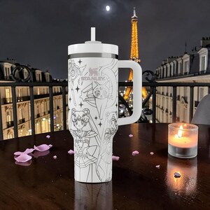 May include: A white Stanley travel mug with a handle and straw, decorated with black and white anime character designs. The mug sits on a wooden table with a lit candle and rose petals, with the Eiffel Tower visible.