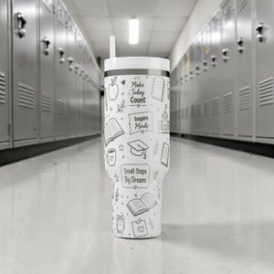 May include: A white tumbler with a black rim and a straw. The tumbler is decorated with black line drawings of an apple, books, a graduation cap, and the phrases "Make Today Count", "Inspire Minds", and "Small Steps Big Dreams".