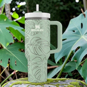 Puede incluir: Un vaso Stanley verde con tapa y asa blancas, con un dise&ntilde;o de hojas tropicales. Tiene una pajita y el logotipo de Stanley. El fondo incluye hojas verdes grandes y una roca cubierta de musgo.