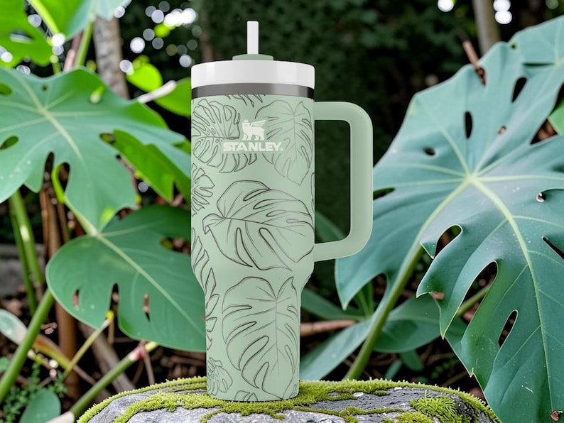 Puede incluir: Un vaso Stanley verde con tapa y asa blancas, con un dise&ntilde;o de hojas tropicales. Tiene una pajita y el logotipo de Stanley. El fondo incluye hojas verdes grandes y una roca cubierta de musgo.