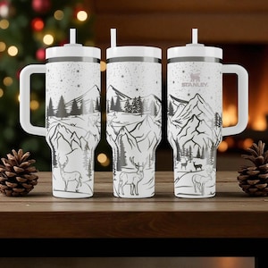 K&ouml;nnte beinhalten: Drei wei&szlig;e isolierte Becher mit schwarzen und grauen Berg- und Hirschmotiven. Jeder Becher hat einen wei&szlig;en Griff, Deckel und Strohhalm. Das Stanley-Logo ist auf einem Becher sichtbar. Die Becher stehen auf einer Holzoberfl&auml;che.