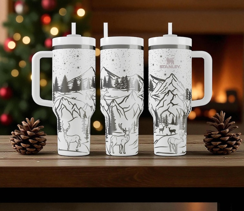 K&ouml;nnte beinhalten: Drei wei&szlig;e isolierte Becher mit schwarzen und grauen Berg- und Hirschmotiven. Jeder Becher hat einen wei&szlig;en Griff, Deckel und Strohhalm. Das Stanley-Logo ist auf einem Becher sichtbar. Die Becher stehen auf einer Holzoberfl&auml;che.