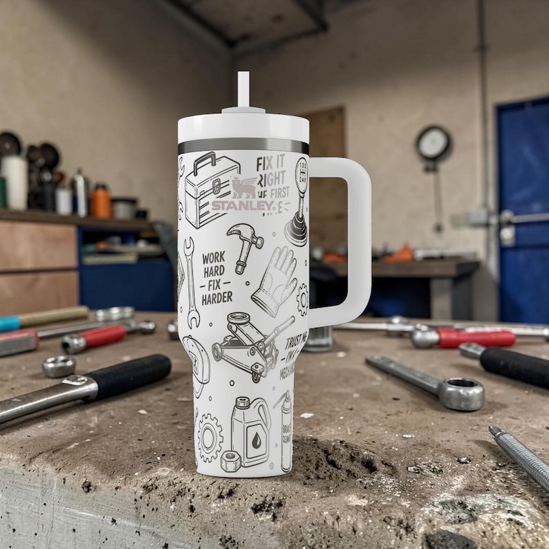 May include: White insulated tumbler with a handle and a straw. The tumbler is decorated with black line drawings of tools, a toolbox, and the text "Fix it right, Work hard, Fix harder, Trust the process". The tumbler is sitting on a workbench.