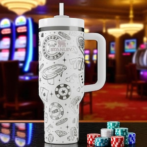 May include: A white Stanley tumbler with a black casino-themed design, including roulette wheels, poker chips, and playing cards. The tumbler has a white handle and lid with a straw. A collection of poker chips in various colors is in the foreground.