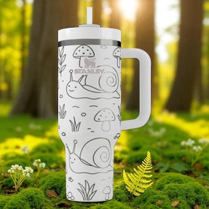 May include: White Stanley tumbler with a handle, featuring a whimsical design of snails, mushrooms, and foliage. The tumbler has a white lid and a straw. The Stanley logo is visible. The background is a blurred forest scene.