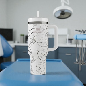 May include: A white Stanley tumbler with a handle and straw, featuring a repeating black line art pattern of lips, syringes, and vials. The Stanley logo is printed in black on the upper portion of the cup, which has a cylindrical shape.