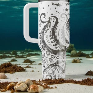 May include: A white travel mug with a large handle, featuring a black and white octopus design. The design includes tentacles, fish, and other marine elements. The mug is set against a blurred underwater background.