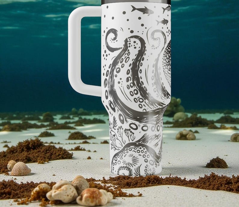 May include: A white travel mug with a large handle, featuring a black and white octopus design. The design includes tentacles, fish, and other marine elements. The mug is set against a blurred underwater background.