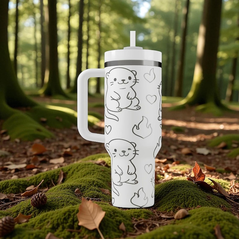 K&ouml;nnte beinhalten: Wei&szlig;er Becher mit Griff und Strohhalm, verziert mit einem sich wiederholenden Muster aus Otter-Illustrationen und Herzen. Der Becher befindet sich vor einem nat&uuml;rlichen Waldhintergrund mit Moos und Herbstlaub.