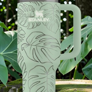 Puede incluir: Un termo Stanley verde claro con un asa grande, con un patr&oacute;n repetido de hojas tropicales. El logotipo de Stanley es visible. El termo est&aacute; rodeado de exuberante follaje verde.