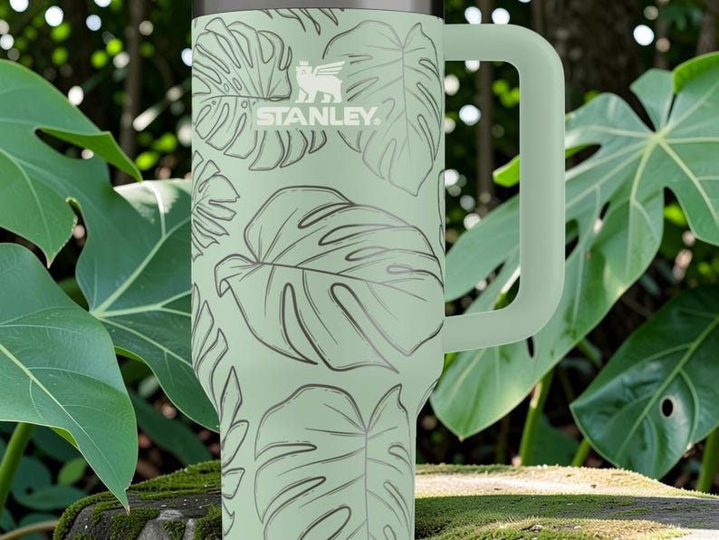 Puede incluir: Un termo Stanley verde claro con un asa grande, con un patr&oacute;n repetido de hojas tropicales. El logotipo de Stanley es visible. El termo est&aacute; rodeado de exuberante follaje verde.