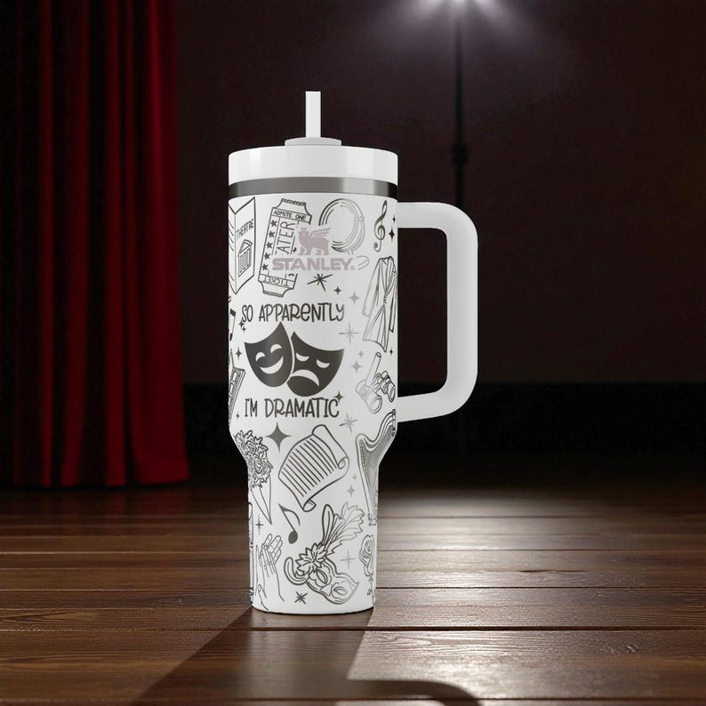 May include: White tumbler with a handle and a white lid, featuring a black and white theatrical design. The design includes drama masks, musical notes, and the text "SO APPARENTLY I'M DRAMATIC."