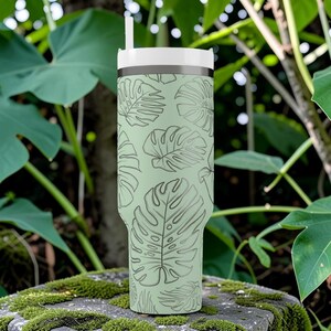 Puede incluir: Un vaso de color verde claro con tapa y pajita blancas, adornado con un patr&oacute;n repetido de hojas de monstera. El vaso es cil&iacute;ndrico y parece ser de acero inoxidable, perfecto para bebidas fr&iacute;as.