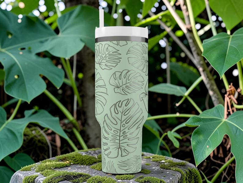 Puede incluir: Un vaso de color verde claro con tapa y pajita blancas, adornado con un patr&oacute;n repetido de hojas de monstera. El vaso es cil&iacute;ndrico y parece ser de acero inoxidable, perfecto para bebidas fr&iacute;as.