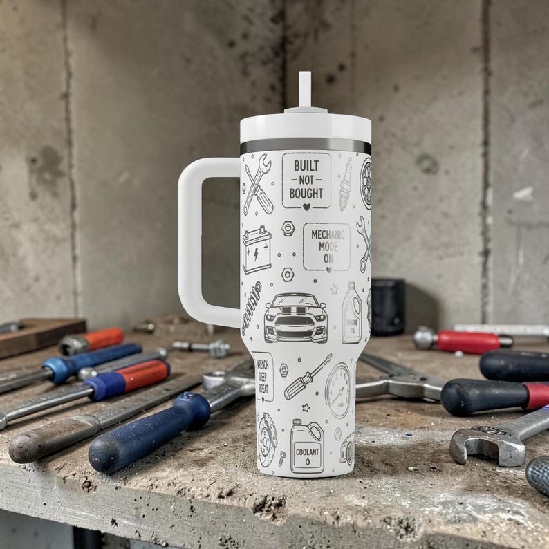 Op de afbeelding: Witte reismok met een handvat en een rietje, met een zwart-wit ontwerp met een mechanisch thema. Het ontwerp bevat gereedschap, een auto en de tekst "BUILT -NOT- BOUGHT" en "MECHANIC MODE ON".
