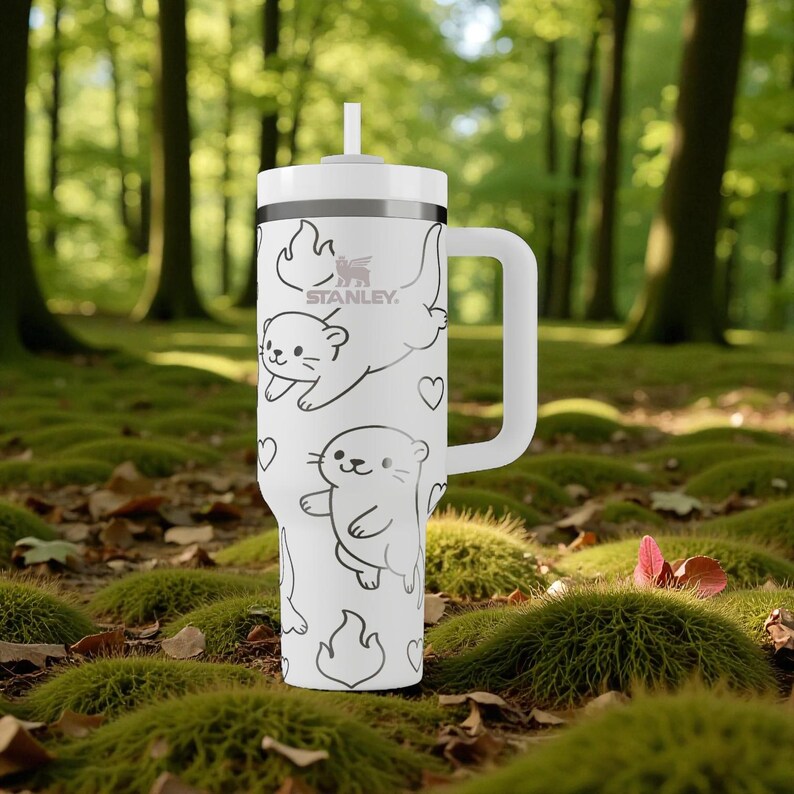 K&ouml;nnte beinhalten: Wei&szlig;er Stanley-Becher mit Griff und Strohhalm. Der Becher ist mit schwarzen Strichzeichnungen von Ottern, Herzen und Flammen verziert. Das Stanley-Logo ist auf der Seite sichtbar. Der Becher steht vor dem Hintergrund eines Waldbodens.
