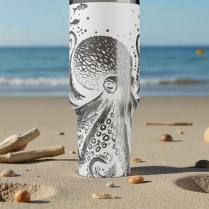 May include: A white travel mug with a black and grey octopus illustration. The design shows the octopus's head, tentacles, and suckers. The mug has a silver rim and a black lid. The background shows a beach with sand and the sea.