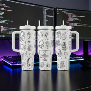 May include: Three white insulated tumblers with black gaming-themed graphics, including headphones, controllers, and keyboards. Each tumbler has a white handle and straw. Text includes "GAME ON", "LEVEL UP", "EAT SLEEP GAME REPEAT", "STAY WIRED", and "GG".