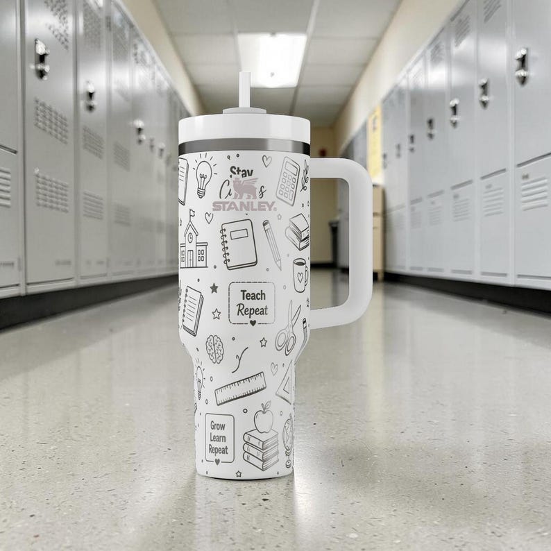 May include: A white Stanley tumbler with a handle and straw, decorated with black line drawings of school supplies and the text "Teach Repeat" and "Grow Learn Repeat". The tumbler is in a school hallway.