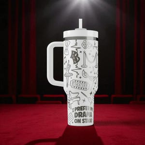 May include: White travel mug with a handle and a straw, featuring a black and white theater-themed design. The design includes bow ties, musical notes, a ballerina, and the text "I PREFER MY DRAMA ON STAGE."
