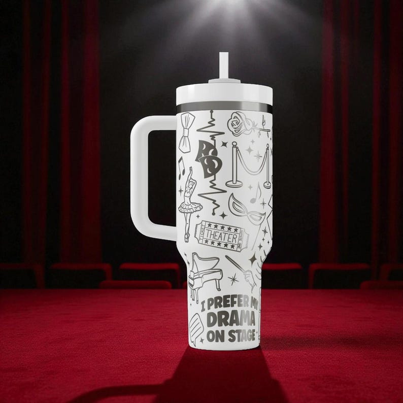 May include: White travel mug with a handle and a straw, featuring a black and white theater-themed design. The design includes bow ties, musical notes, a ballerina, and the text "I PREFER MY DRAMA ON STAGE."