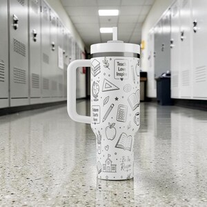 May include: A white insulated tumbler with a handle and a straw. The tumbler is decorated with black line drawings of school-related items, including books, apples, and pencils. The text "Teach Love Inspire" and "Future Starts Here" are also visible.