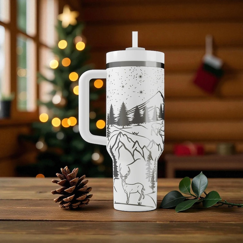 K&ouml;nnte beinhalten: Wei&szlig;er Reisebecher mit einem schwarz-wei&szlig;en Berg- und Hirschdesign. Der Becher hat einen wei&szlig;en Griff, Deckel und Strohhalm. Das Design zeigt eine Bergkette, B&auml;ume und einen Hirsch unter einem Sternenhimmel. Der Becher steht auf einer Holzoberfl&auml;che.