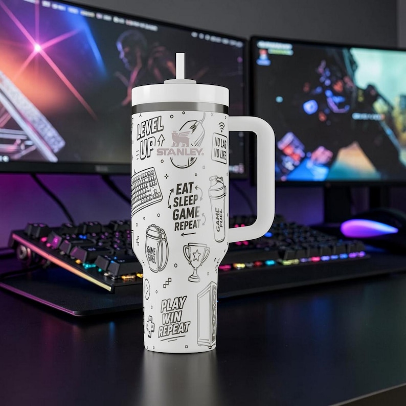 May include: White Stanley tumbler with a handle and straw, featuring black and white gaming-themed graphics. The design includes phrases like "Level Up," "Eat Sleep Game Repeat," and "Play Win Repeat," along with gaming icons. The tumbler is designed for gamers.