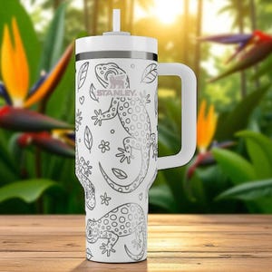 Puede incluir: Vaso Stanley blanco con asa, tapa y pajita. El vaso está decorado con un diseño en blanco y negro de lagartos, hojas y flores. El logotipo de Stanley es visible en el lateral.