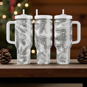 May include: Three white Stanley tumblers with a gray pine needle and pine cone design. Each tumbler has a white handle, lid, and straw. The tumblers are on a wooden surface with pine cones in front. The Stanley logo is visible on one tumbler.