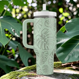 Puede incluir: Un vaso t&eacute;rmico verde claro con asa y tapa blanca. El vaso presenta un patr&oacute;n repetido de hojas de monstera en un verde m&aacute;s oscuro. El fondo es un entorno natural con hojas verdes grandes.