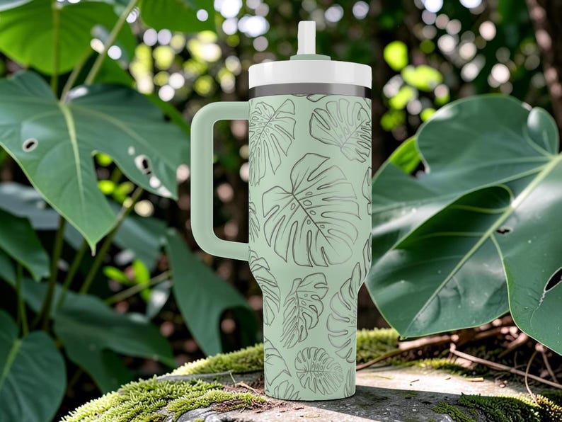 Puede incluir: Un vaso t&eacute;rmico verde claro con asa y tapa blanca. El vaso presenta un patr&oacute;n repetido de hojas de monstera en un verde m&aacute;s oscuro. El fondo es un entorno natural con hojas verdes grandes.