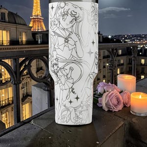 May include: A white tumbler with a black and white manga illustration. The design features a character, stars, and other figures. The tumbler is set against a night cityscape with lit buildings and candles, creating a romantic atmosphere.