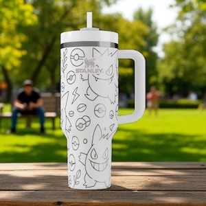 May include: White Stanley tumbler with a handle and straw, featuring a black and white cartoon design of various characters and symbols. The tumbler has a silver band and the Stanley logo. The background is a blurred outdoor scene.