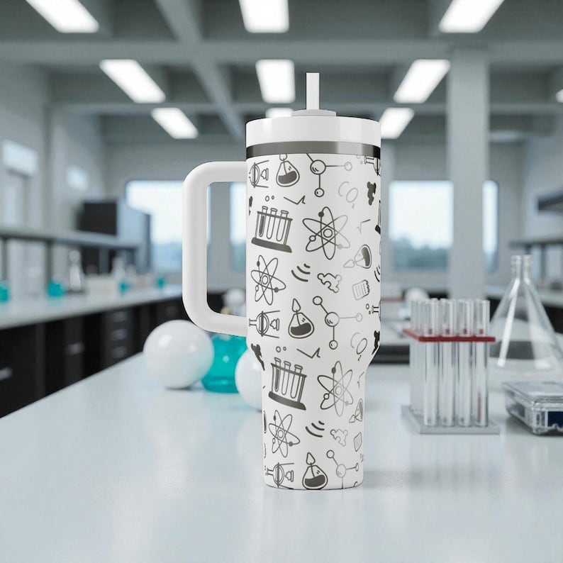May include: A white insulated tumbler with a handle and a straw, decorated with a repeating pattern of science-themed doodles in black. The tumbler is on a white surface in a laboratory setting.