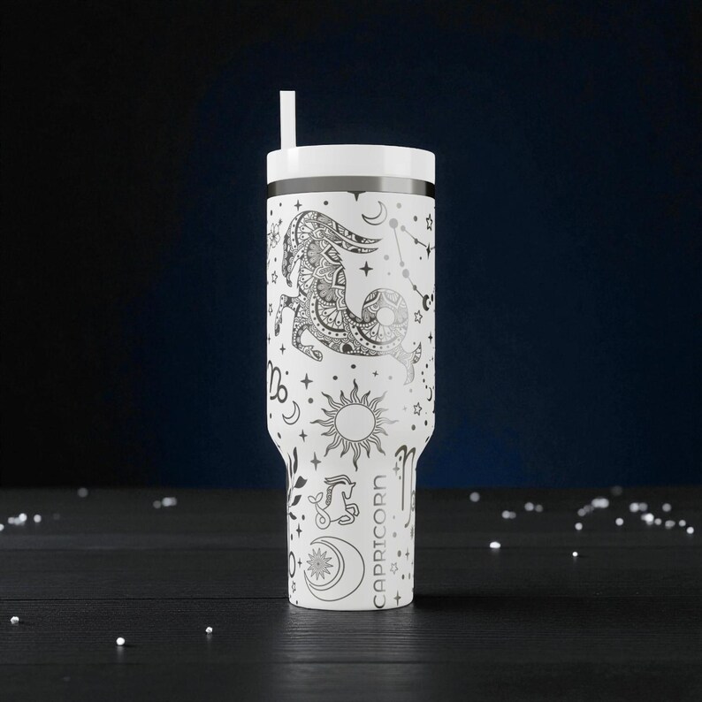 K&ouml;nnte beinhalten: Wei&szlig;er Becher mit Deckel und Strohhalm, mit einem Sternzeichen-Design f&uuml;r Steinbock. Das Design umfasst eine detaillierte Ziegenillustration, Sternbilder, Sonne, Mond und das Wort "STEINBOCK". Der Becher ist etwa 23 cm hoch.