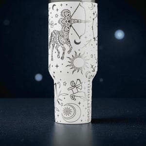 Op de afbeelding: Witte beker met een zwart Boogschutter dierenriem ontwerp. Het ontwerp bevat een centaur boogschutter, sterrenbeelden, zon, maan en sterren. Het woord "SAGITTARIUS" staat verticaal op de zijkant.