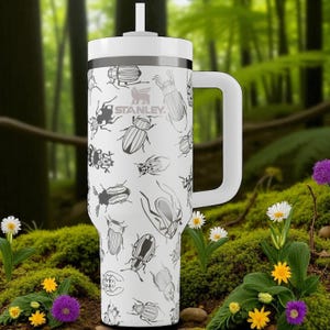 Könnte beinhalten: Weißer Stanley-Becher mit einem schwarzen Käfermuster, einem weißen Griff und einem weißen Deckel. Das Stanley-Logo ist sichtbar. Der Becher befindet sich vor einem verschwommenen Hintergrund mit Wald, Blumen und Moos.