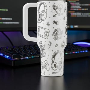 May include: White travel mug with a large white handle, featuring a black and white gaming-themed design. The design includes headphones, game controllers, and text that reads "GAME ON" and "JUST ONE MORE GAME".