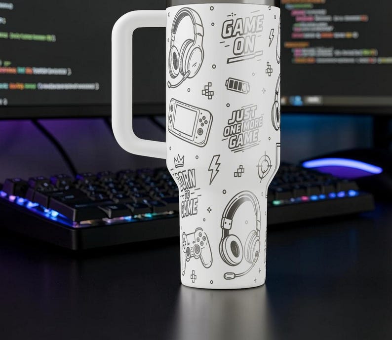 May include: White travel mug with a large white handle, featuring a black and white gaming-themed design. The design includes headphones, game controllers, and text that reads "GAME ON" and "JUST ONE MORE GAME".