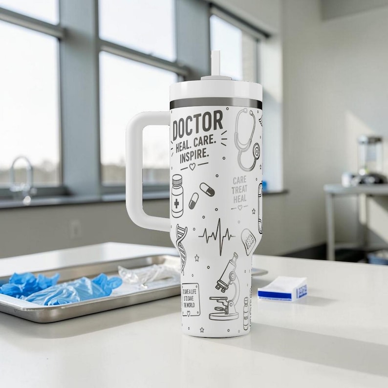 May include: White insulated tumbler with a handle and a straw. The tumbler is decorated with black line art of medical symbols, including a stethoscope, pills, and a DNA strand. Text on the tumbler reads "DOCTOR HEAL. CARE. INSPIRE."